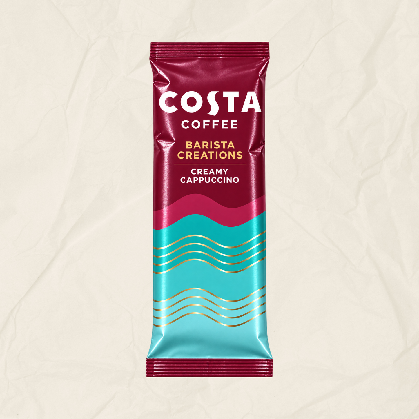 A single Costa Coffee Barista Creations Creamy Cappuccino sachet standing upright on a textured beige background. The packaging features a deep magenta top half with white branding, a bright turquoise bottom half with gold wavy lines, and a magenta-to-turquoise gradient center.