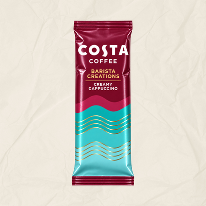A single Costa Coffee Barista Creations Creamy Cappuccino sachet standing upright on a textured beige background. The packaging features a deep magenta top half with white branding, a bright turquoise bottom half with gold wavy lines, and a magenta-to-turquoise gradient center.