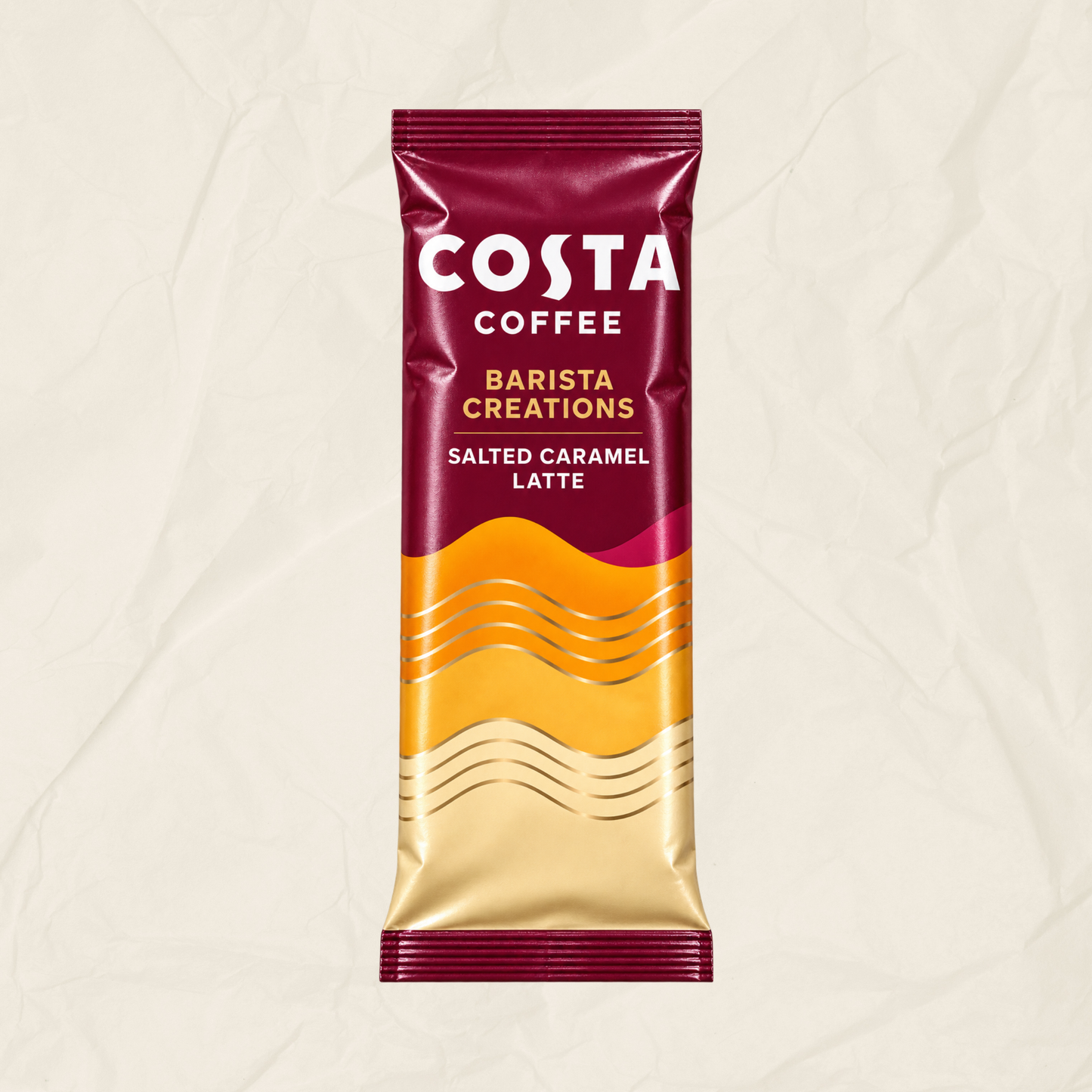 A single Costa Coffee Barista Creations Salted Caramel Latte sachet standing upright on a textured off-white background. The top half is a burgundy colour with white branding, the lower half is a yellow gradient with wavy lines.