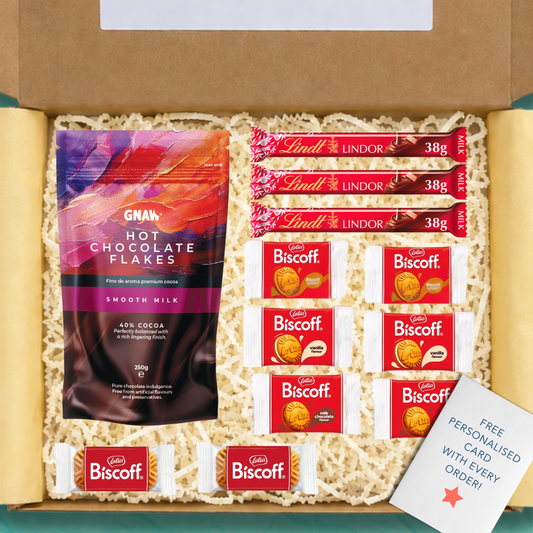 An open gift box with yellow tissue paper and yellow shredded paper packaging containing a series of items: A pack of GNAW hot chocolate flakes, 3 Lindt Lindor chocolate bars, a selection of Lotus Biscoff biscuits, and a free personalised card.