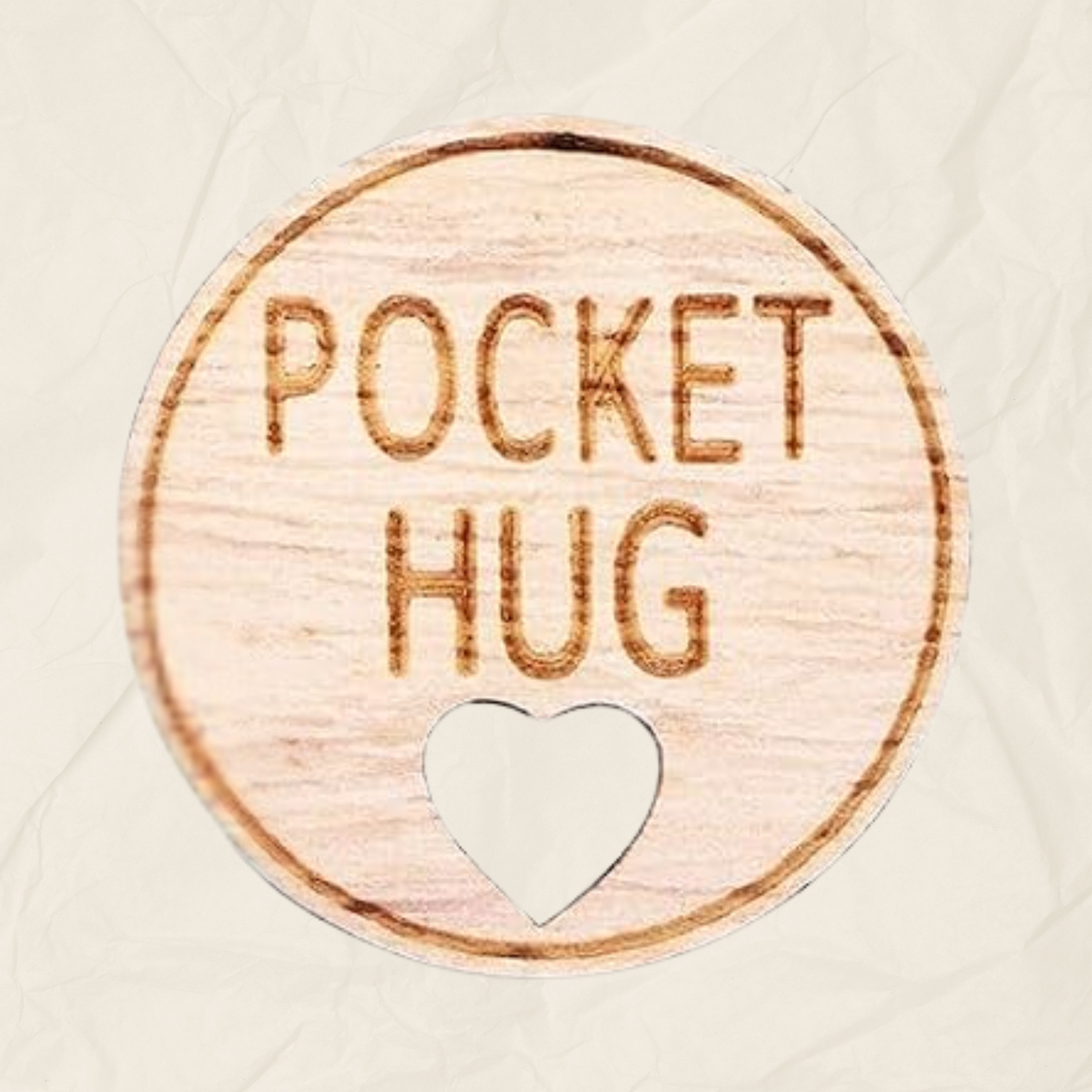 A round wooden token with the text "Pocket Hug" laser etched into it and a heart shaped hole, designed to be kept in the recipients pocket.