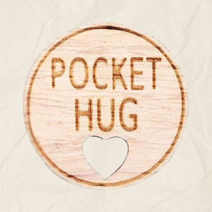 A round wooden token with the text "Pocket Hug" laser etched into it and a heart shaped hole, designed to be kept in the recipients pocket.