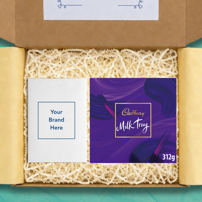 An open gift box containing a purple Cadbury Milk Tray chocolate bar package with the size 312g on the box in white text, and a white personalised card that reads 'Your brand here', surrounded by beige crinkle paper filler on yellow wrapping paper.