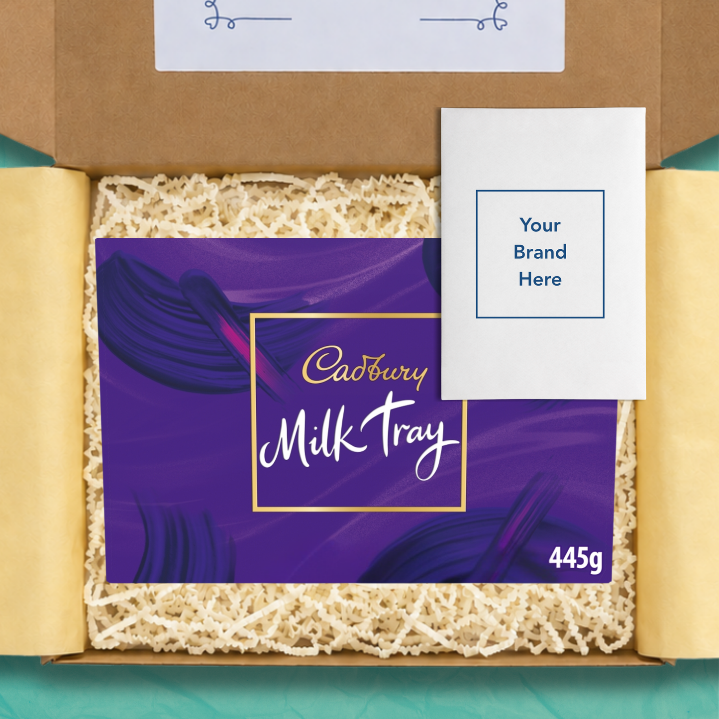An open gift box containing a purple Cadbury Milk Tray chocolate bar package with the size 445g on the box in white text, and a white personalised card that reads 'Your brand here', surrounded by beige crinkle paper filler on yellow wrapping paper.