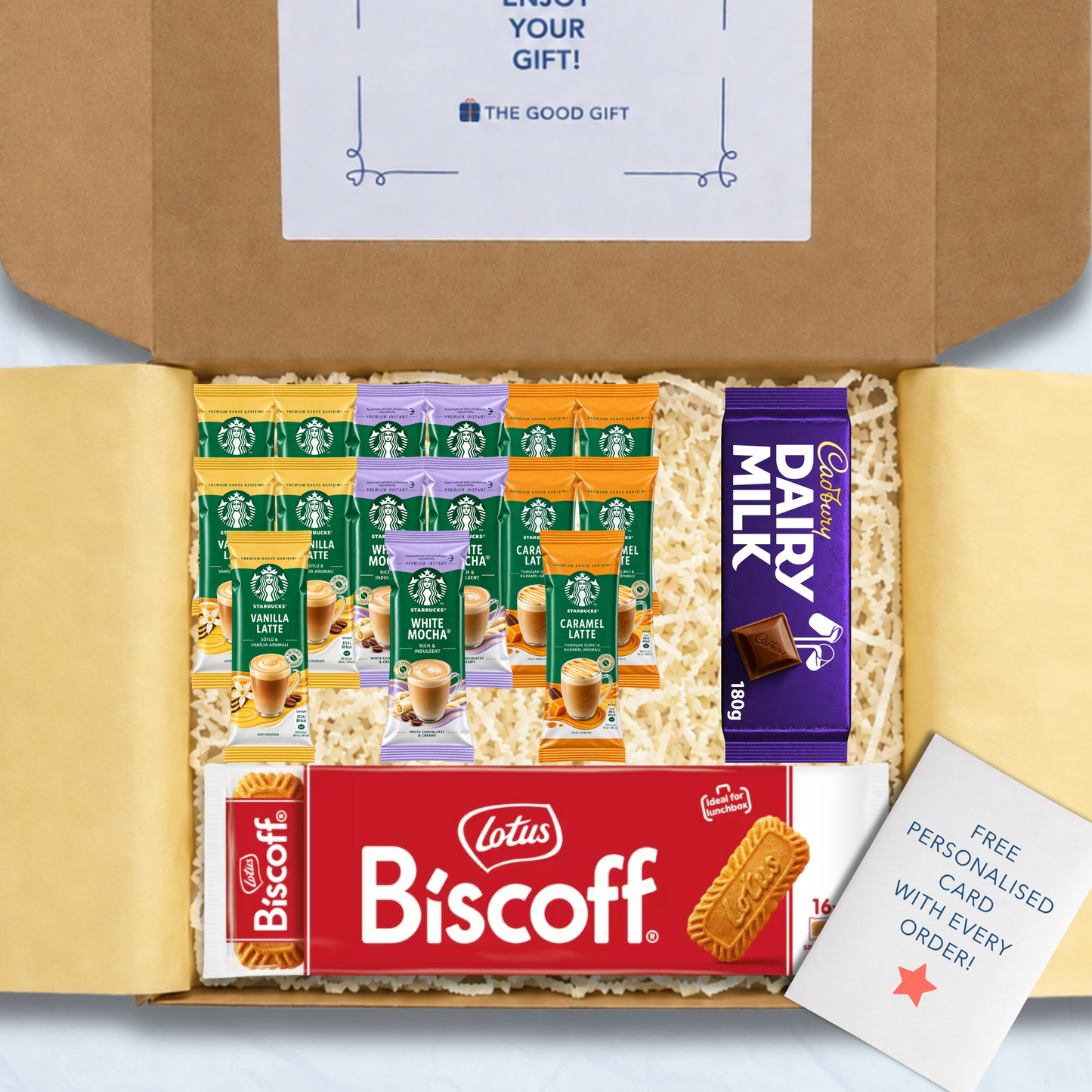 A curated gift box filled with packaged snacks and biscuits, including Starbucks coffee sachets, a pack of Lotus Biscuit, a Cadbury 180g Dairy Milk chocolate bar, and a free personalised card, shown from above inside an open brown cardboard box.