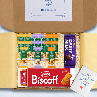 A curated gift box filled with packaged snacks and biscuits, including Starbucks coffee sachets, a pack of Lotus Biscuit, a Cadbury 180g Dairy Milk chocolate bar, and a free personalised card, shown from above inside an open brown cardboard box.
