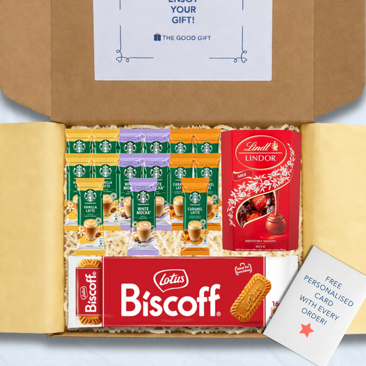 A curated gift box filled with packaged snacks and biscuits, including Starbucks coffee sachets, a pack of Lotus Biscuit, a Lindt Lindor 200g box of milk chocolate truffles, and a free personalised card, shown from above inside an open brown cardboard box.