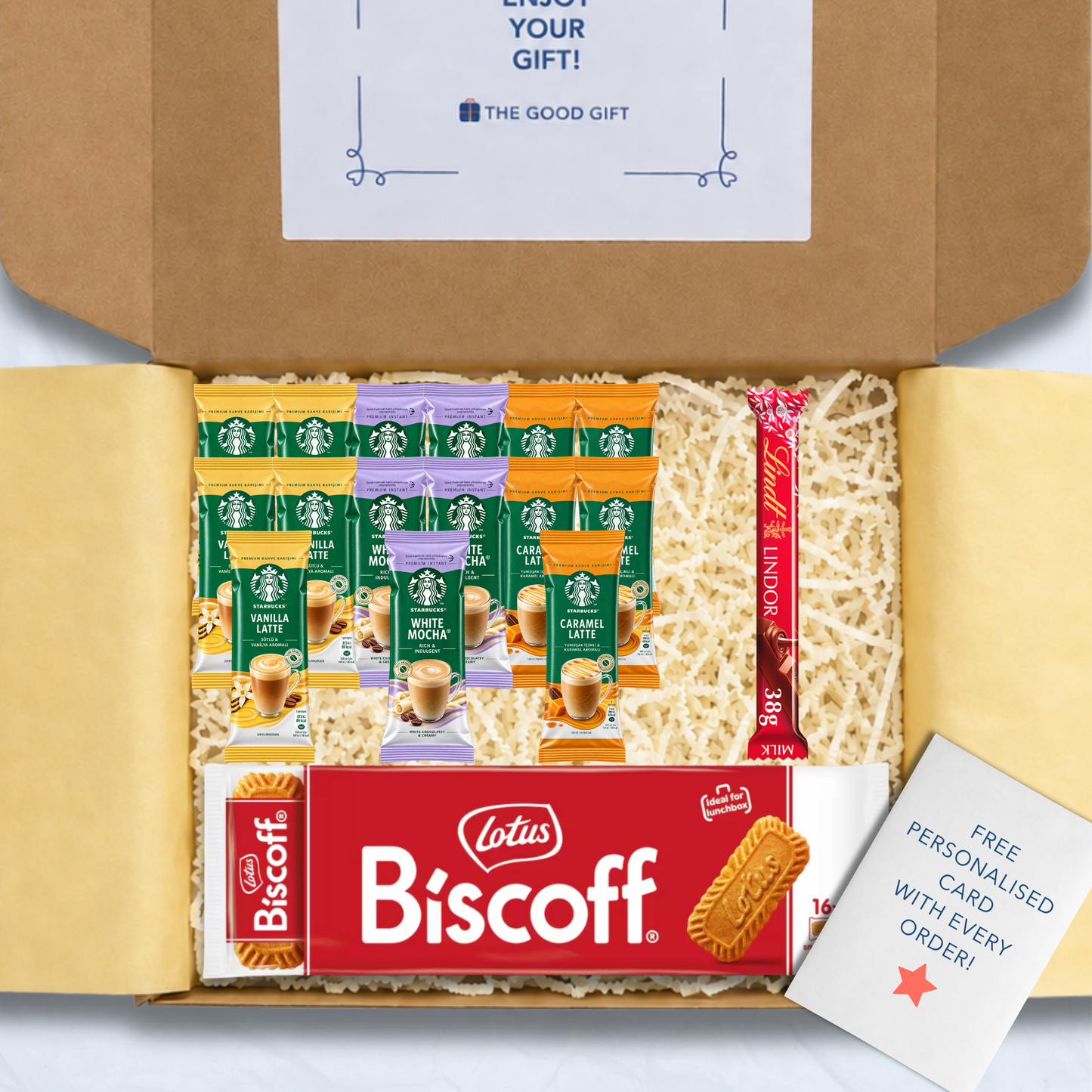 A curated gift box filled with packaged snacks and biscuits, including Starbucks coffee sachets, a pack of Lotus Biscuit, a Lindt Lindor 38g milk chocolate bar, and a free personalised card, shown from above inside an open brown cardboard box.