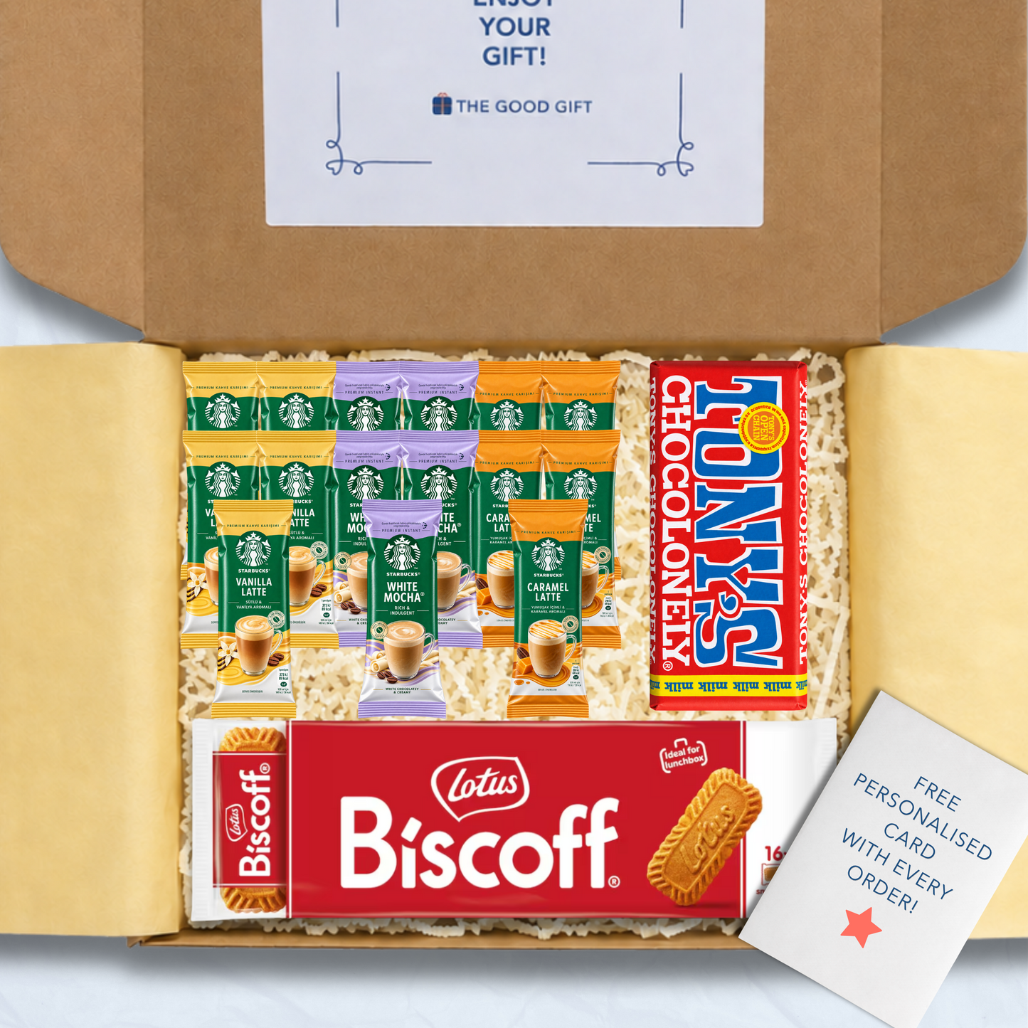 A curated gift box filled with packaged snacks and biscuits, including Starbucks coffee sachets, a pack of Lotus Biscuit, a Tony's Chocolonely milk chocolate bar, and a free personalised card, shown from above inside an open brown cardboard box.