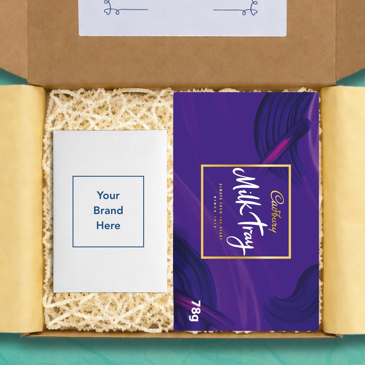 An open gift box containing a purple Cadbury Milk Tray chocolate bar package with the size 78g on the box in white text, and a white personalised card that reads 'Your brand here', surrounded by beige crinkle paper filler on yellow wrapping paper.
