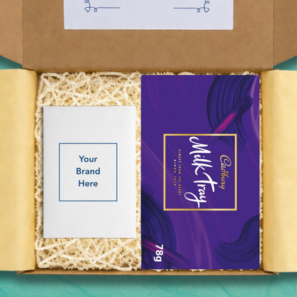An open gift box containing a purple Cadbury Milk Tray chocolate bar package with the size 78g on the box in white text, and a white personalised card that reads 'Your brand here', surrounded by beige crinkle paper filler on yellow wrapping paper.
