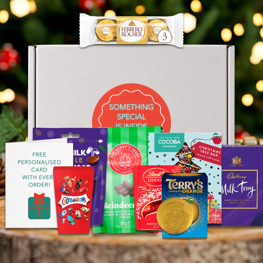 A gift set featuring a white box behind an array of items including: Ferrero Rocher, Lindt Lindor Chocolate Truffles, Celebrations chocolates, Terrys chocolate orange, Cadbury Dairy Milk Tray, Cadbury Little Robins, Cocoba Christmas Tree chocolate bar, a pack of Reindeer Poo chocolate biscuit and caramel pieces, and a chocolate coin, alongside a white personalized card with a green gift icon and teal text. The products are arranged on a rustic wooden surface with a blurred Christmas tree in the background.