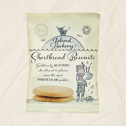 A package of Island Bakery Shortbread Biscuits on a textured off-white background. The packaging is cream-colored with black text and illustrations of a cow, birds, and a kilted figure playing bagpipes, with two biscuits pictured at the bottom.