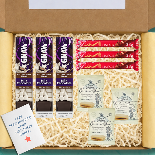 An open gift box with yellow tissue paper and yellow shredded packing paper. The box contains three GNAW hot chocolate spoons, three Lindt Lindor 38g chocolate bars, three packets of Island Bakery shortbread biscuits, and a free personalised card.