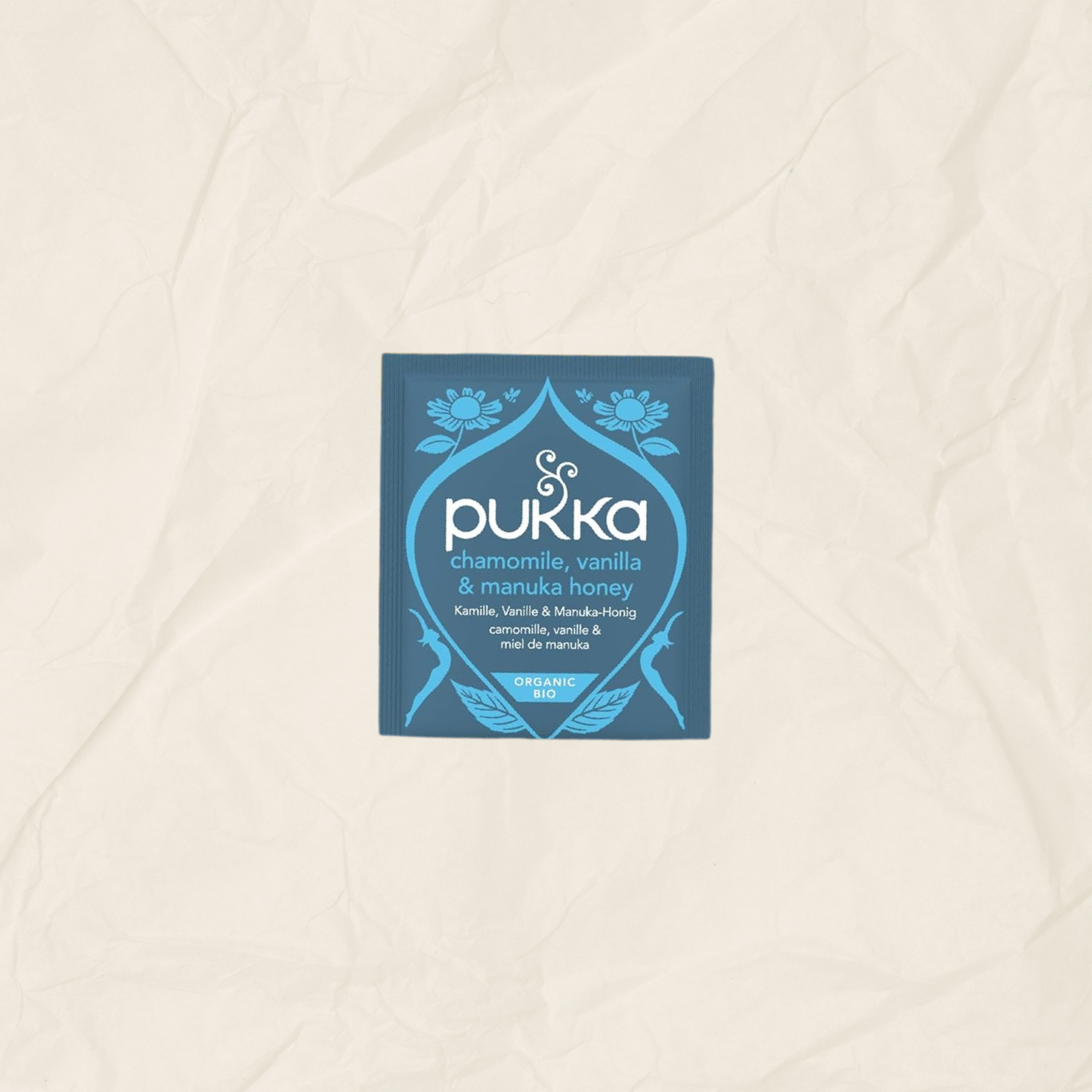 A single square Pukka tea packet in blue and white, labeled chamomile, vanilla & manuka honey, centered on a textured beige paper background.