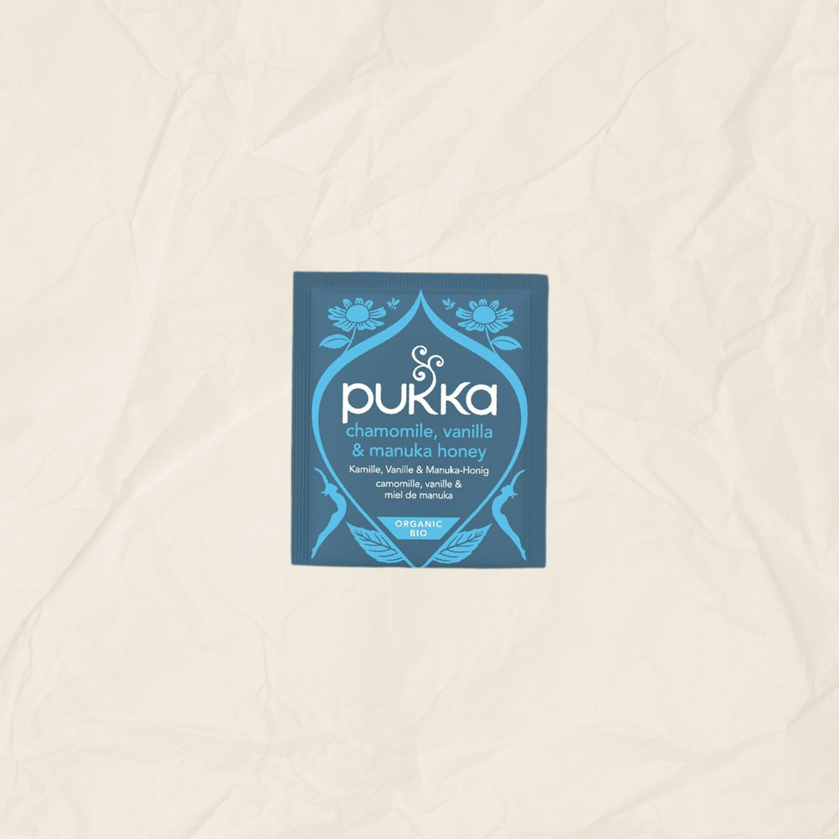 A single square Pukka tea packet in blue and white, labeled chamomile, vanilla & manuka honey, centered on a textured beige paper background.