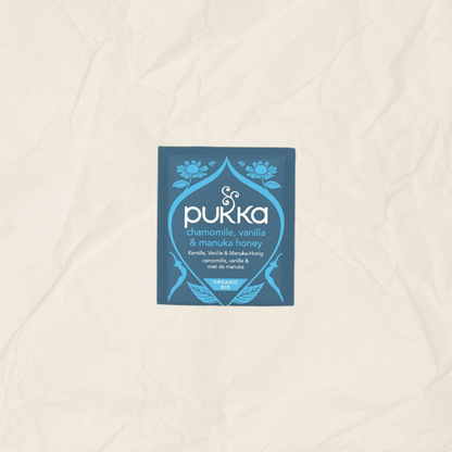 A single square Pukka tea packet in blue and white, labeled chamomile, vanilla & manuka honey, centered on a textured beige paper background.