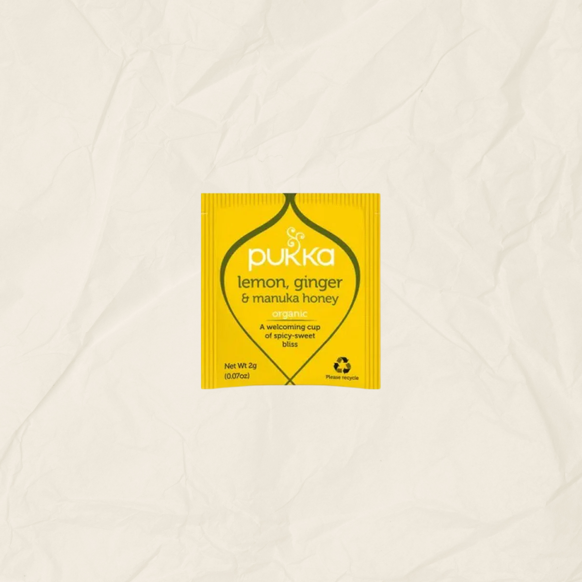 A single yellow Pukka lemon, ginger & manuka honey tea packet centered on a textured, off-white crumpled paper background.