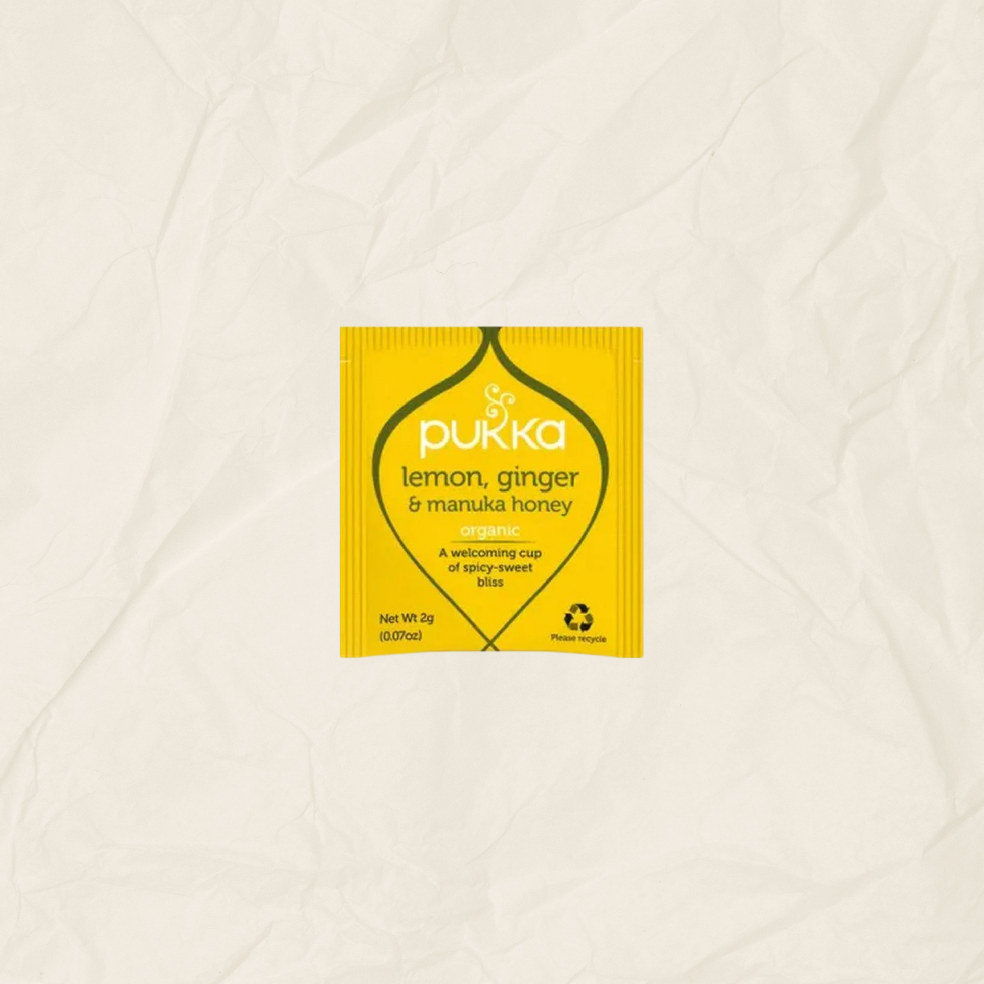 A single yellow Pukka lemon, ginger & manuka honey tea packet centered on a textured, off-white crumpled paper background.