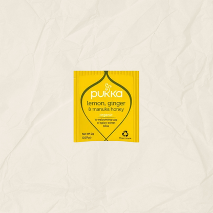 A single yellow Pukka lemon, ginger & manuka honey tea packet centered on a textured, off-white crumpled paper background.