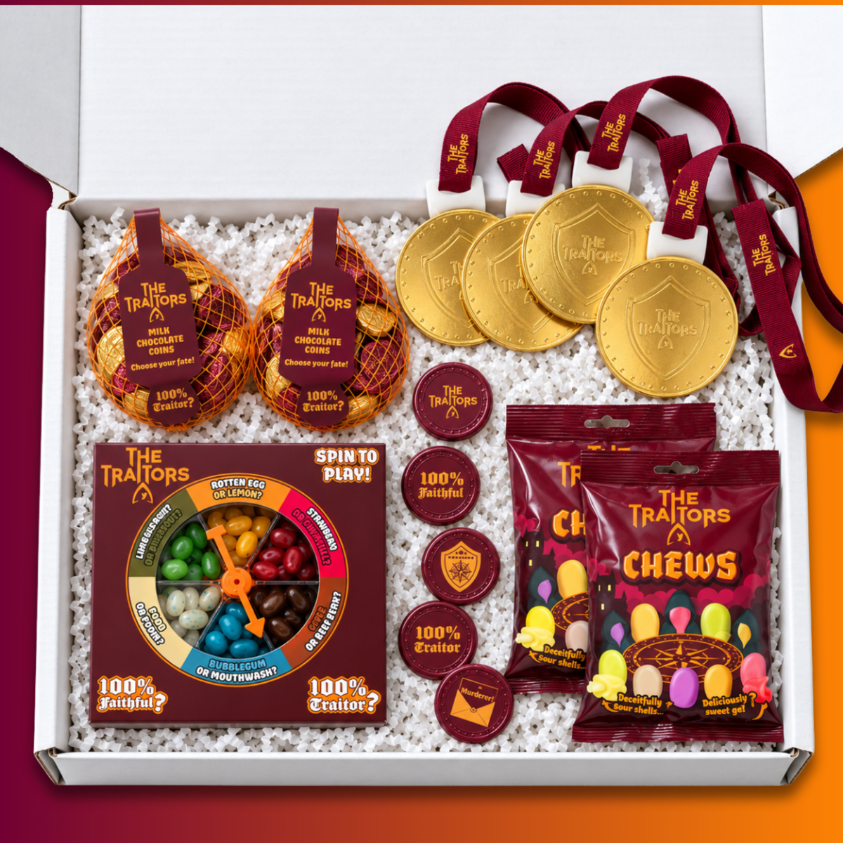 An open gift box filled with assorted candy and branded accessories from The Traitors x Sweets. Visible items include gold-toned chocolate medallion charms with burgundy ribbons, two bags of The Traitors Chews, The Traitors "Spin to win" game, and chocolate coins all arranged on white packing paper.