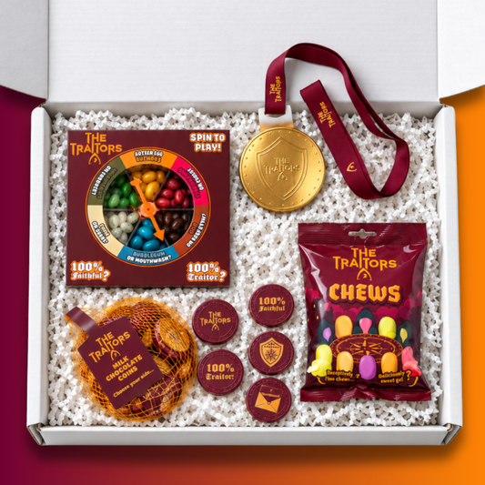 A curated gift box set from The Traitors x Sweets, displayed inside an open white box filled with white crinkle paper. The set includes a gold-tone chocolate medal with a burgundy ribbon, a The Traitors "Spin to win" game, a bag of The Traitors Chews and a bag of chocolate coins.