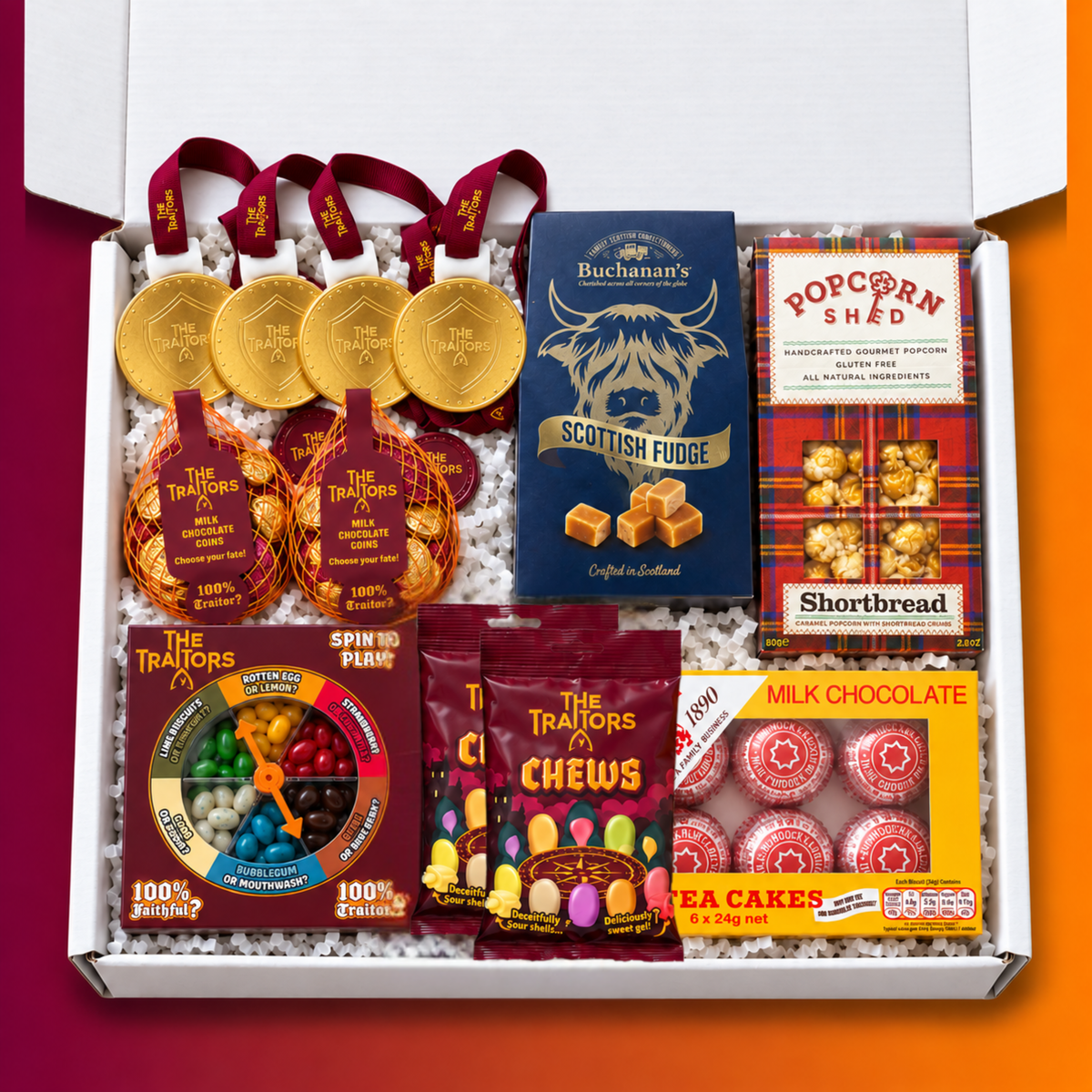 A curated gift box of The Traitors snacks and an assortment of Scottish treats, neatly arranged in white crinkle paper inside an open white box. The assortment includes gold foil-wrapped chocolate medallions, a blue packet of Buchanan's Scottish Fudge, a red-and-black patterned packet of Popcorn Shed Shortbread popcorn, a yellow packet of Tea Cakes, a bag of The Traitors Chews, 2 bags of chocolate coins, and a Traitors "Spin to win" game.