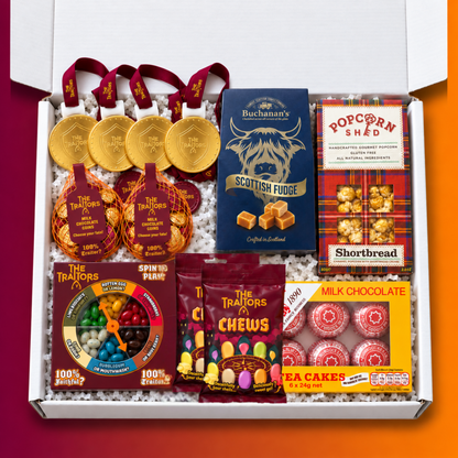 A curated gift box of The Traitors snacks and an assortment of Scottish treats, neatly arranged in white crinkle paper inside an open white box. The assortment includes gold foil-wrapped chocolate medallions, a blue packet of Buchanan's Scottish Fudge, a red-and-black patterned packet of Popcorn Shed Shortbread popcorn, a yellow packet of Tea Cakes, a bag of The Traitors Chews, 2 bags of chocolate coins, and a Traitors "Spin to win" game.